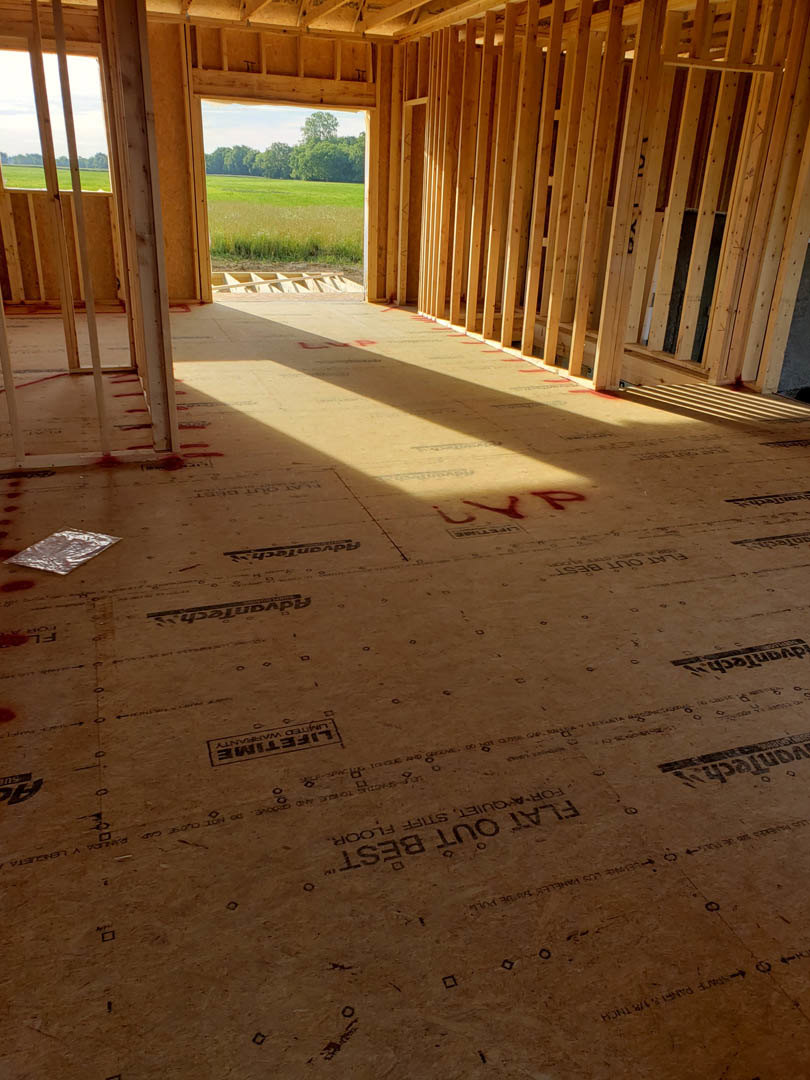 Wood-framed room under construction with exposed beams, unfinished wooden walls, and subfloor featuring handwritten black markings, overlooking grassy field through open framing.
