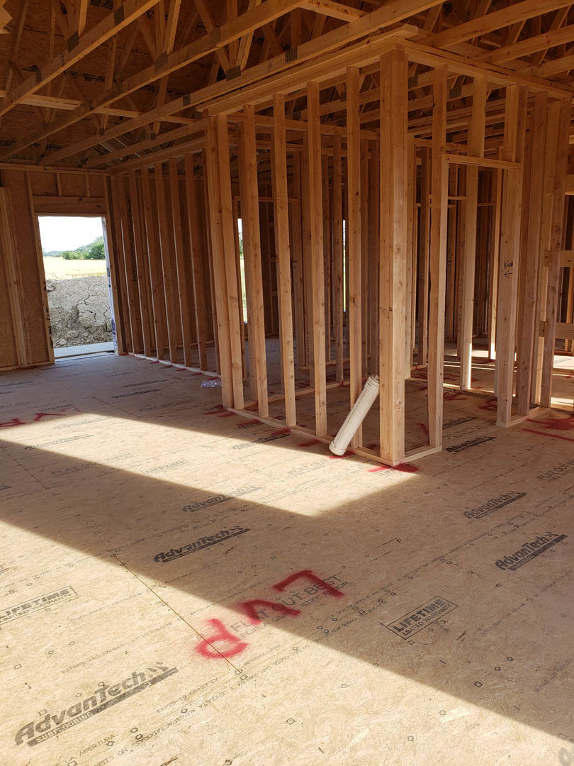 Wood-framed house under construction with exposed beams, white pipe on subfloor, red markings on paper and floor, dry land visible through open doorway