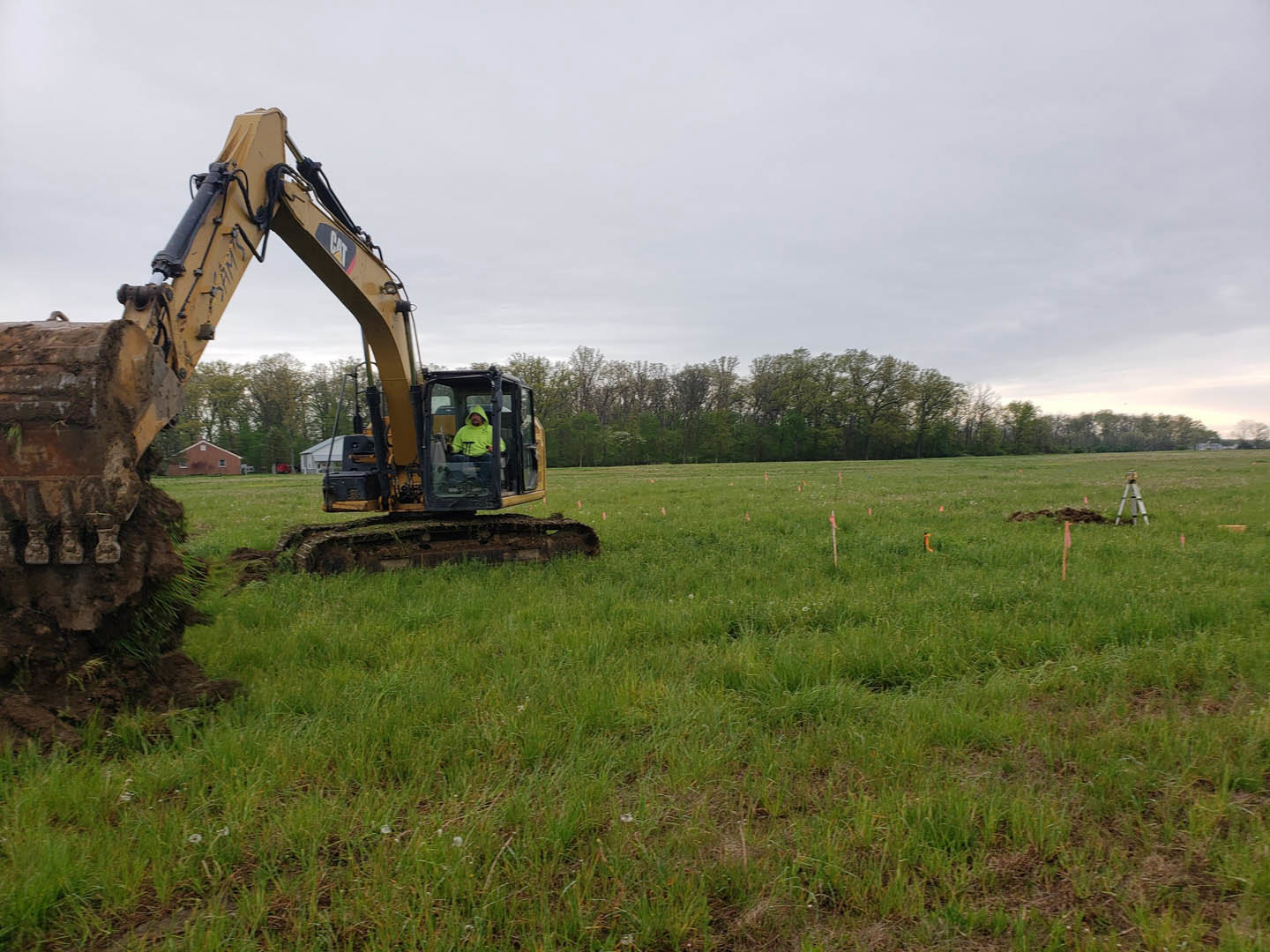 Bulldozer digging in grassy field with scattered trees, cloudy sky overhead, construction workers nearby