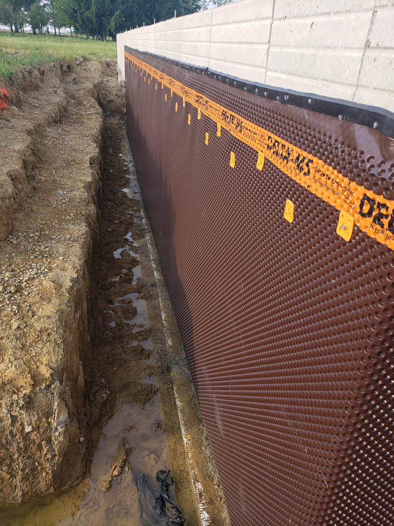 Brown exterior wall with multiple circular holes, orange plastic sheeting partially covering surface, white building and green trees visible in background.