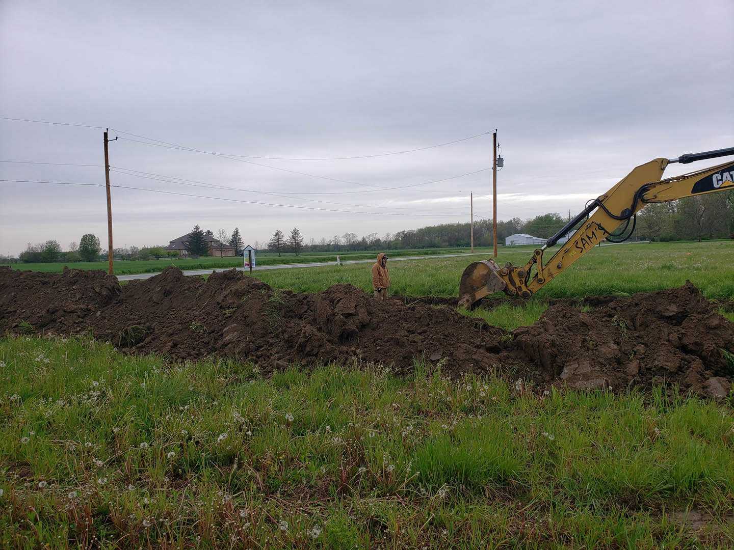 Yellow excavator digging soil in grassy field, man in brown jacket standing nearby, pile of dirt, power lines and trees in background, cloudy sky overhead.