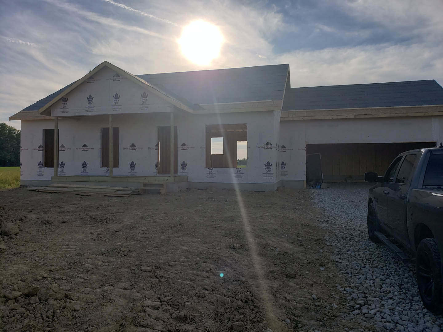 Modern home under construction with exposed framing, unfinished exterior walls, and large windows; parked car in driveway; bright sunlight illuminating site.