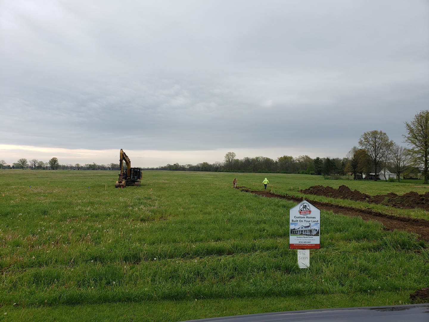 Grassy field with a yellow tractor, a sign posted on the lawn, leafless tree, and cloudy sky overhead