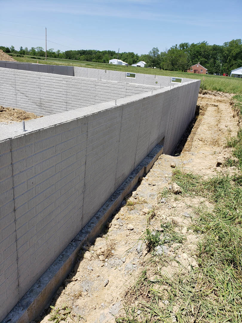 Concrete exterior wall with circular opening in the ground, surrounded by dirt and grass under a partly cloudy blue sky