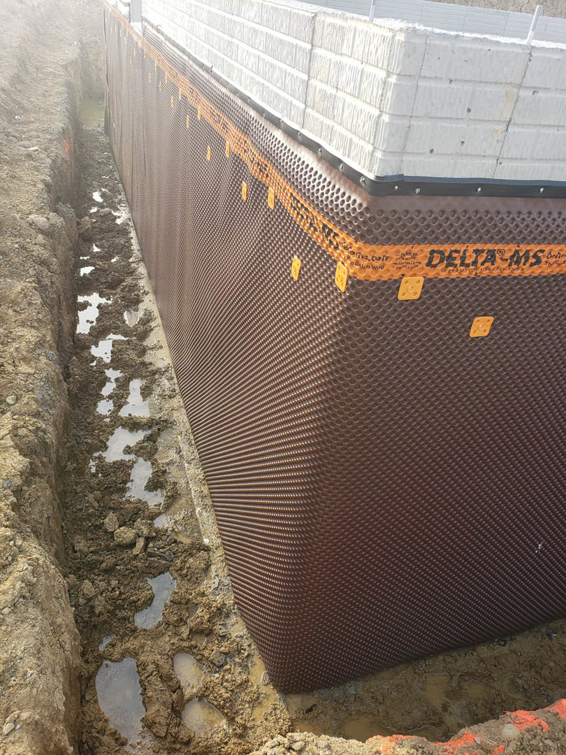 Close-up of an exterior composite wall with textured black and orange surface adjacent to muddy ground