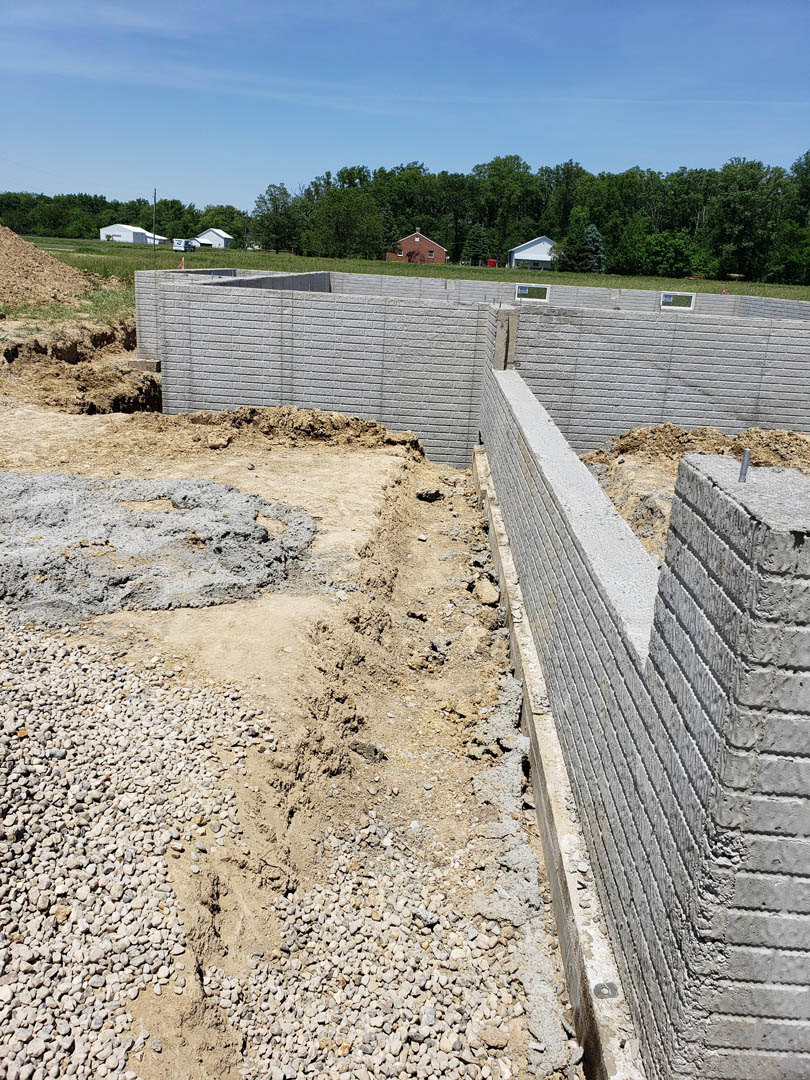 Brick wall under construction with exposed concrete foundation, piles of gravel and dirt in foreground, blue sky and forested trees in background