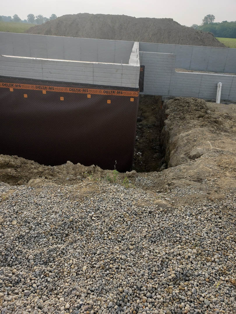 Excavated construction site with exposed dirt and rocks, black and orange tarp covering part of the ground, grassy area and sky in background