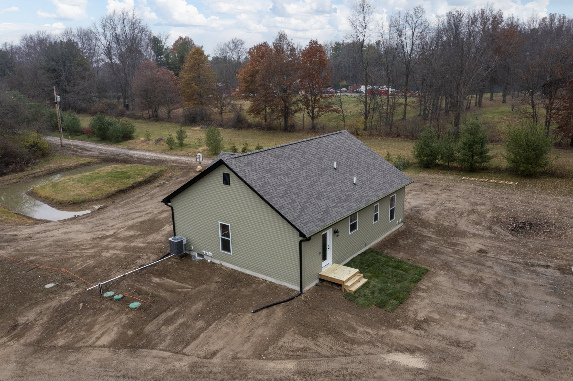 Single-story home with light siding, black roof, and wooden steps leading to the entrance, situated on a dirt lot with scattered brown-leaved trees and a black air conditioner unit