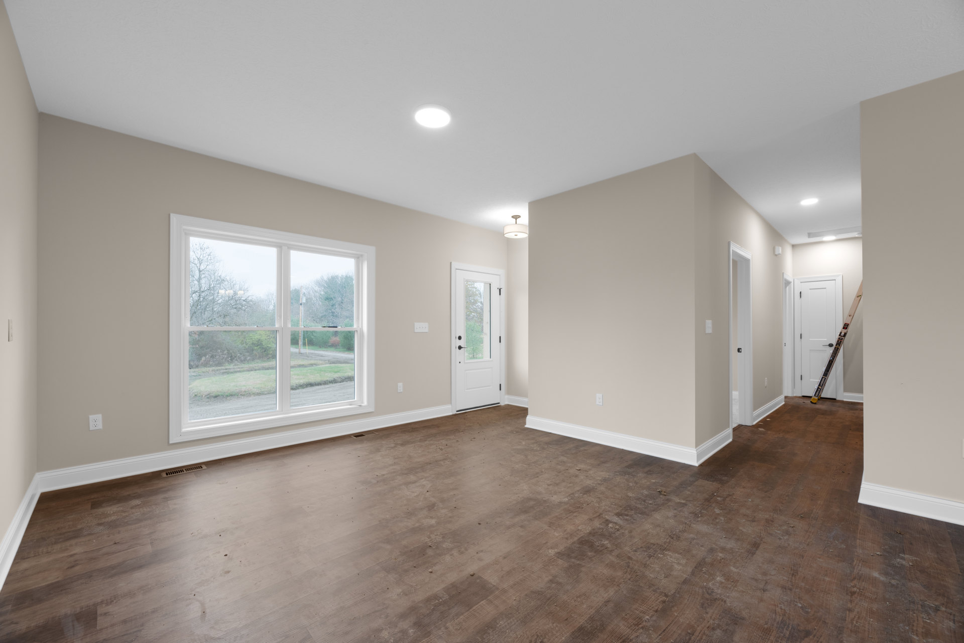 White walls and wood laminate flooring, ceiling light fixture, window overlooking road and trees, white door with glass panel, white baseboard trim, ladder positioned near door