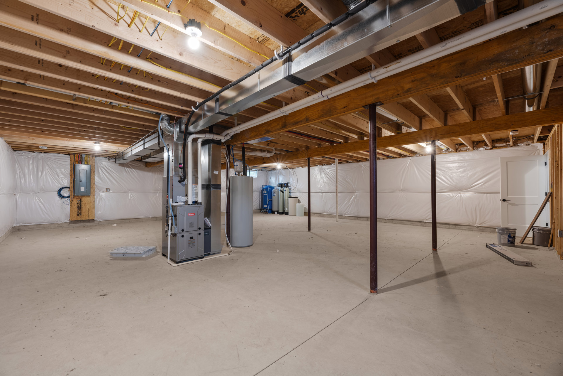 Spacious room featuring exposed steel beams, large cylindrical metal structure, white walls, visible metal pipes, grey bucket on concrete floor, single light bulb with black cover