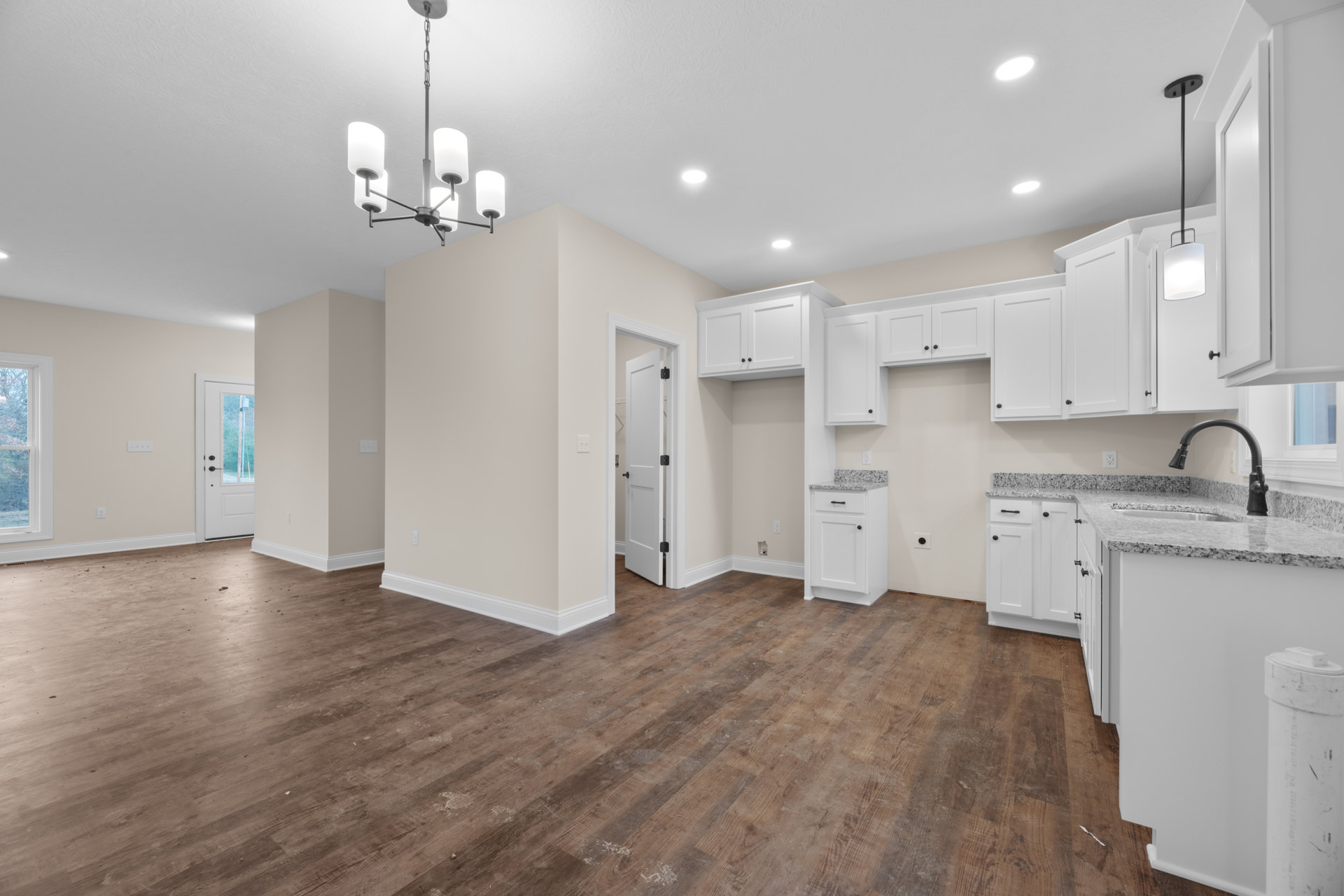 Kitchen with white shaker cabinets, wood laminate flooring, white framed window, black hardware on white door, recessed ceiling lights, and light stone countertops.