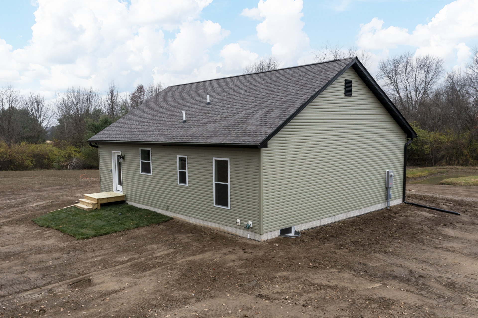 Modern cottage with gray roof, large windows, wooden steps leading to a dirt path, surrounded by trees and clouds in the sky