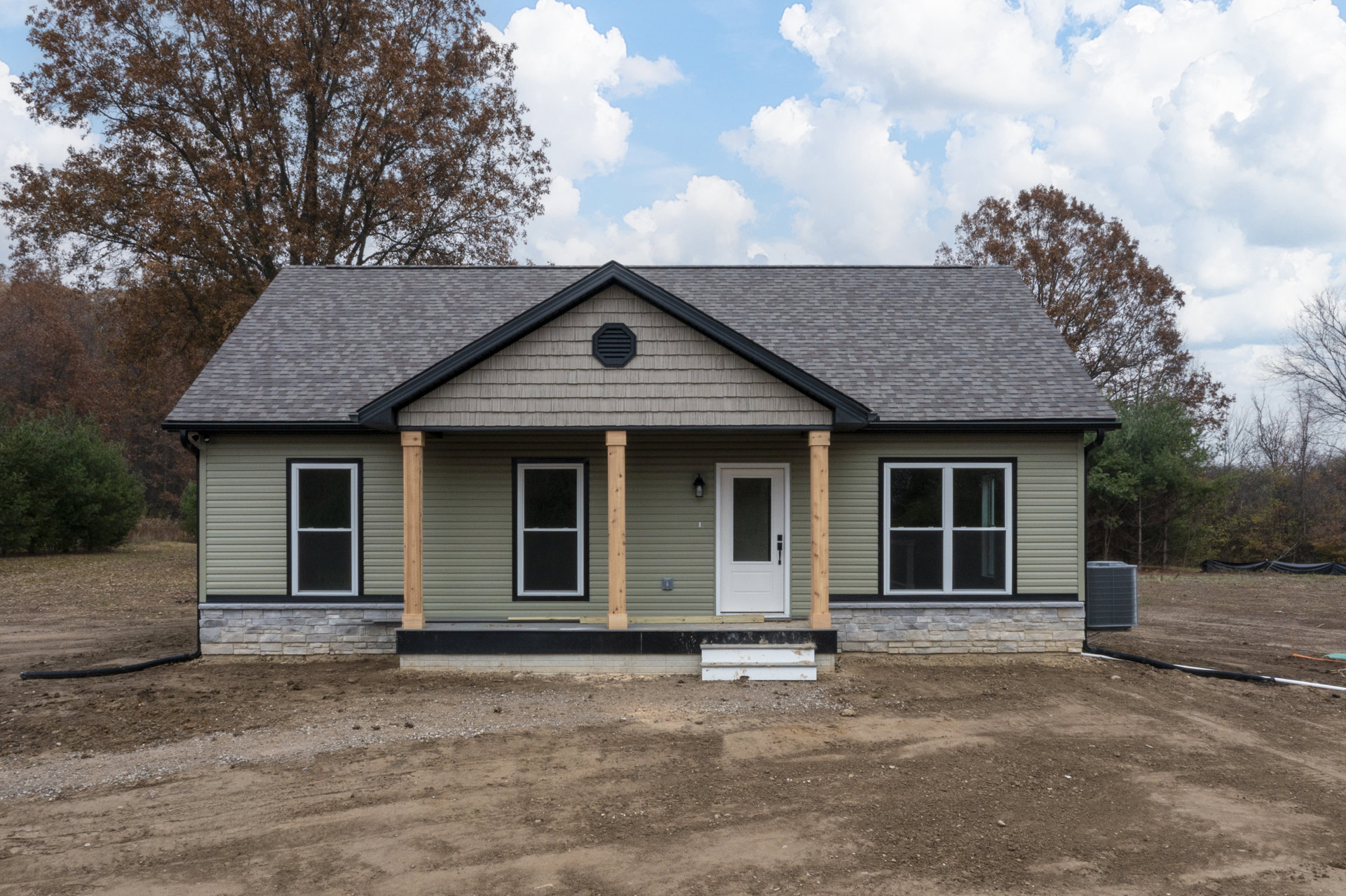 White cottage-style home with white-framed windows, glass-paneled door, and wall vent, set along a dirt road under a cloudy sky.