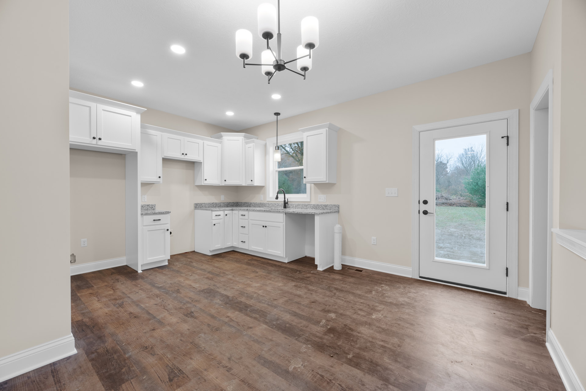 White kitchen cabinets, wood flooring, chandelier with white lamps, door with window overlooking yard and trees, wall-mounted white light fixture