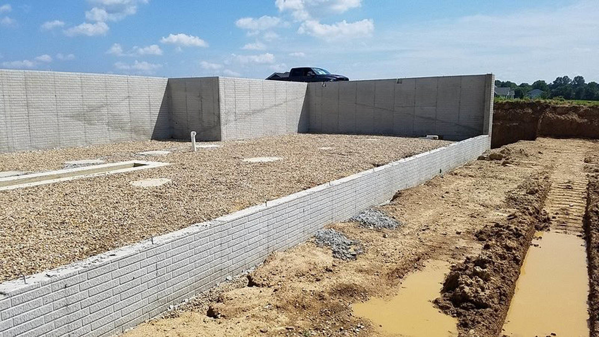Partially built brick home with exposed foundation, white exterior wall, black roof, and construction dirt in foreground; parked car visible in background.