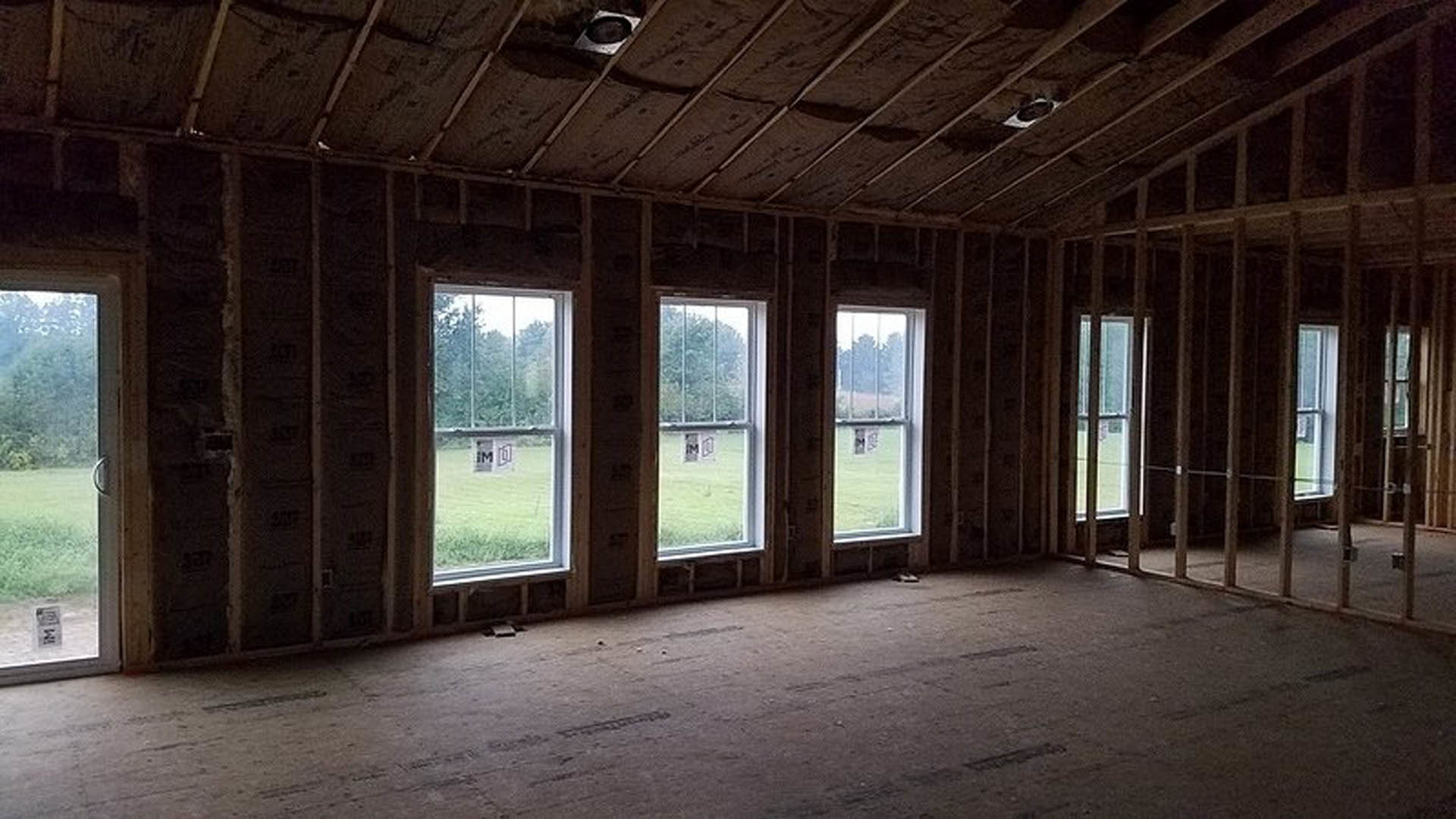 Sunlit room featuring large windows, light wood flooring, exposed ceiling beam with built-in speaker, and neutral walls.