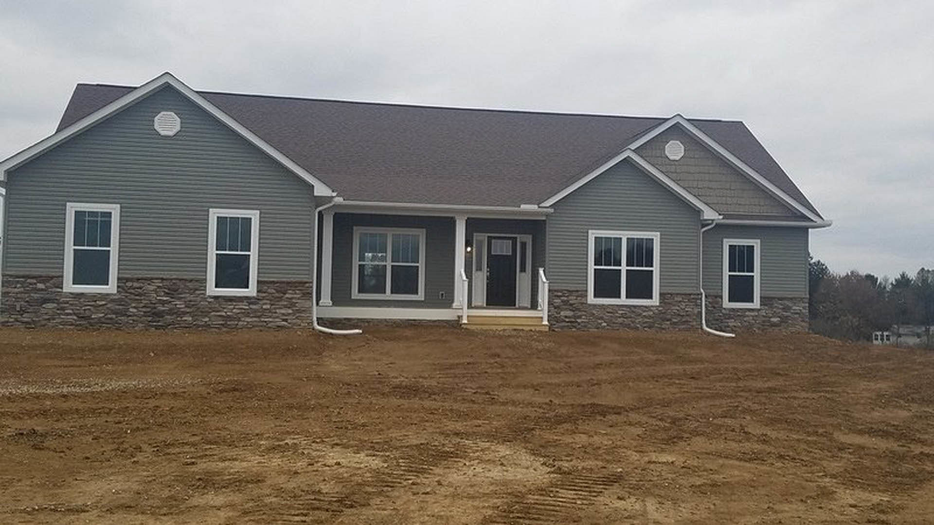 Gray-sided house with black front door, white-framed windows, covered porch, and unfinished dirt yard under cloudy sky