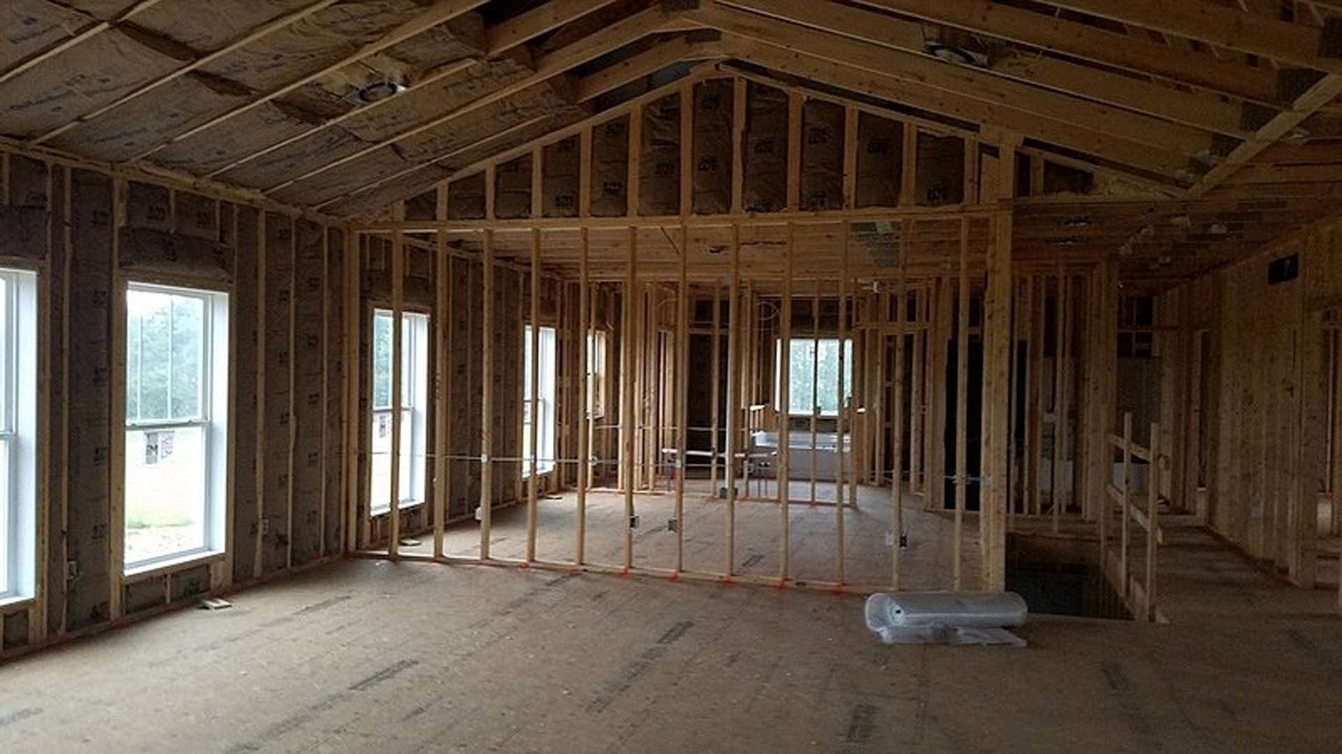 Room under construction with exposed wood framing, white-framed window with glass pane, unfinished wood floor, black square electrical outlet on wall, natural daylight illuminating