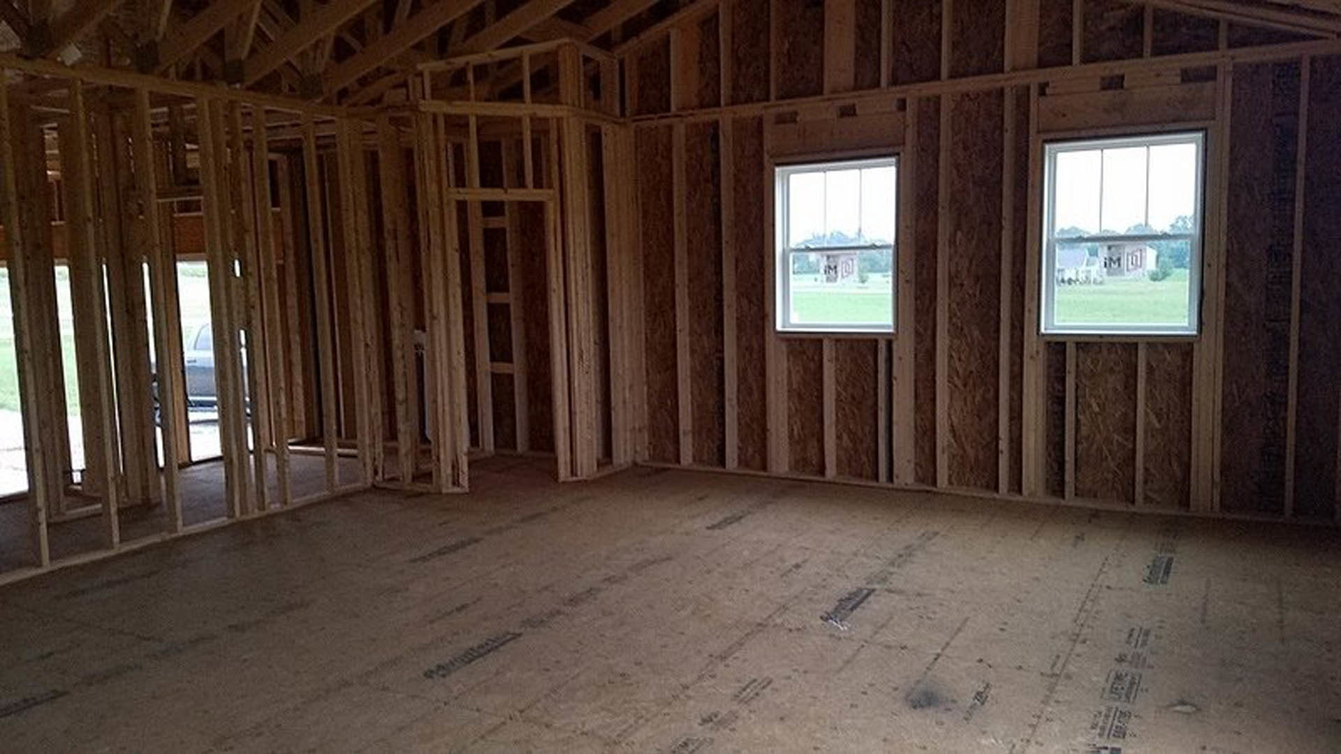 Room under construction with exposed wood framing, unfinished plank flooring, large windows showing exterior building and sign
