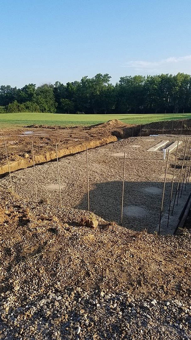 Concrete foundation slab surrounded by dirt and metal poles at a construction site, with grassy land and trees in the background under a partly cloudy blue sky