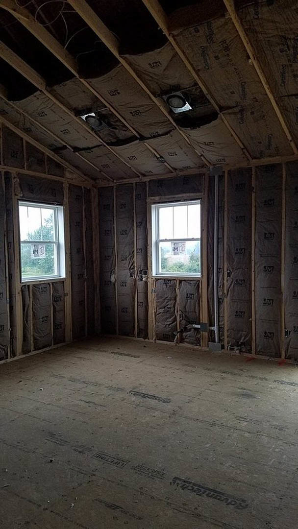 Spacious room with light wood flooring, multiple large windows, white walls, exposed ceiling beams, and a ceiling-mounted light fixture; one window features a sign.