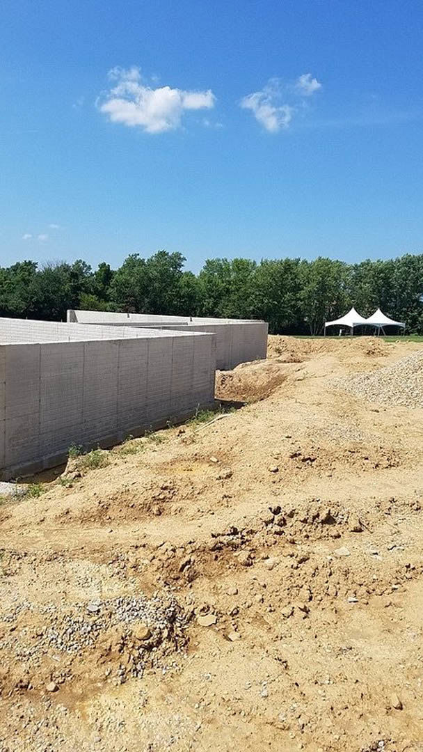 Large mound of dirt and gravel beside a partially constructed concrete wall, with scattered concrete blocks, white exterior wall, and trees in the background.