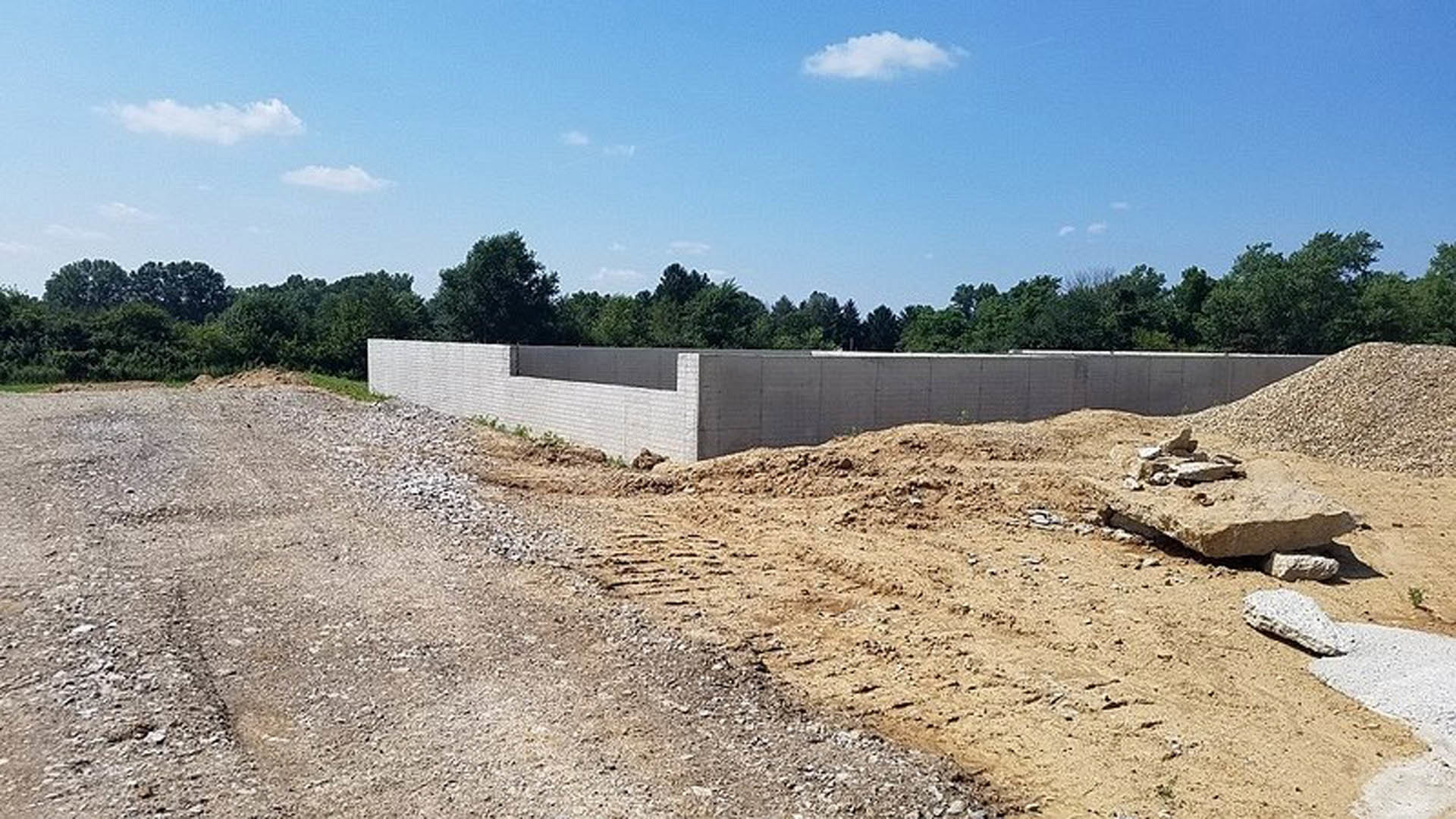 Concrete foundation wall surrounded by exposed soil, construction rubble, and mature trees under a partly cloudy sky