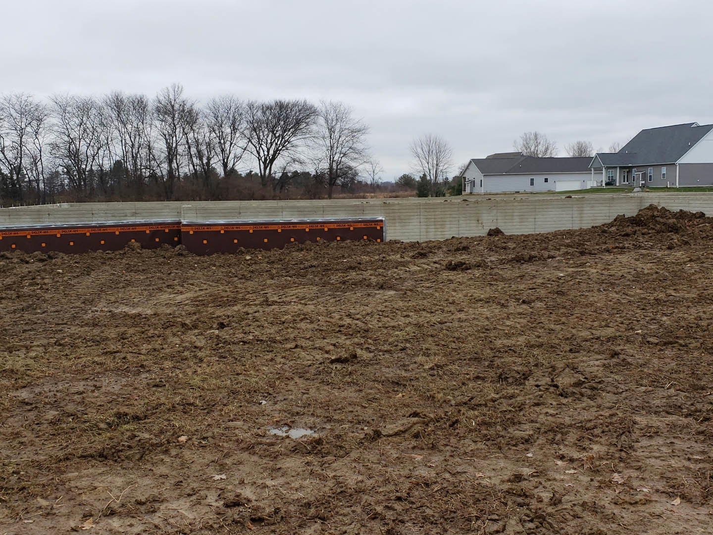 Dirt field bordered by a row of trees, custom home with light-colored siding and attached garage visible in the background