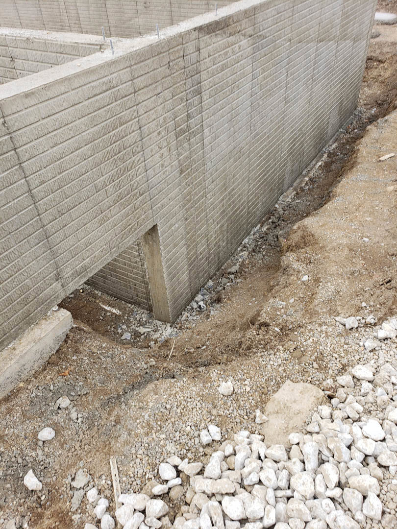 Concrete wall with exposed aggregate, rough ground surface featuring a hole, scattered rocks and rubble, outdoor setting