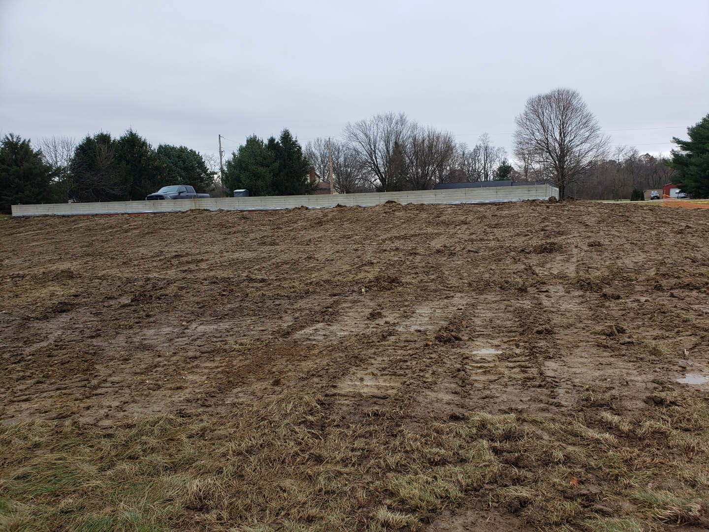 Dirt field bordered by trees, parked car in the background, open sky above, soil and grass covering ground