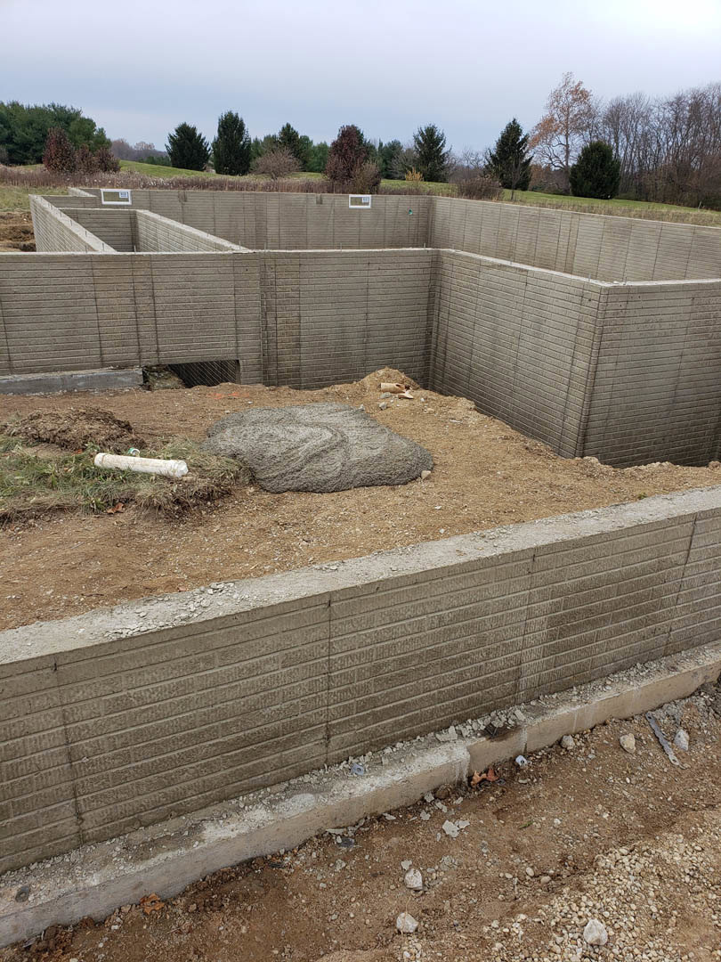 Concrete foundation wall with circular opening in soil, surrounded by grassy field, distant trees, and hay bale under open sky