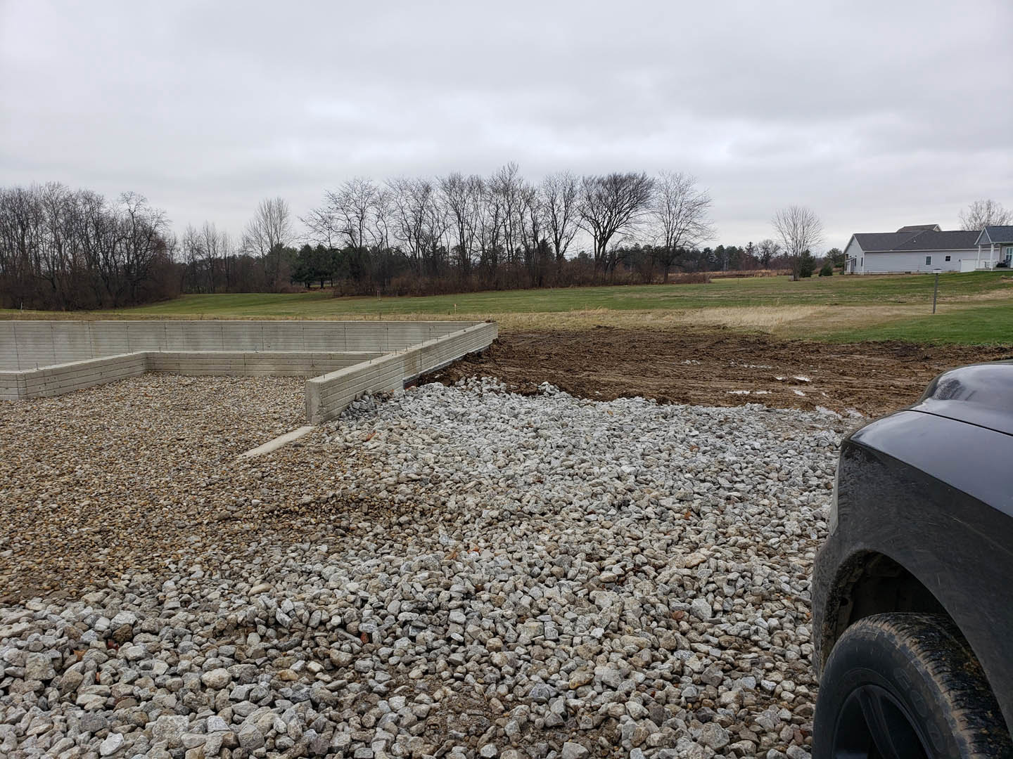 White house with grey roof, concrete wall, gravel and grass foreground, parked car with visible tire, leafless trees in background.