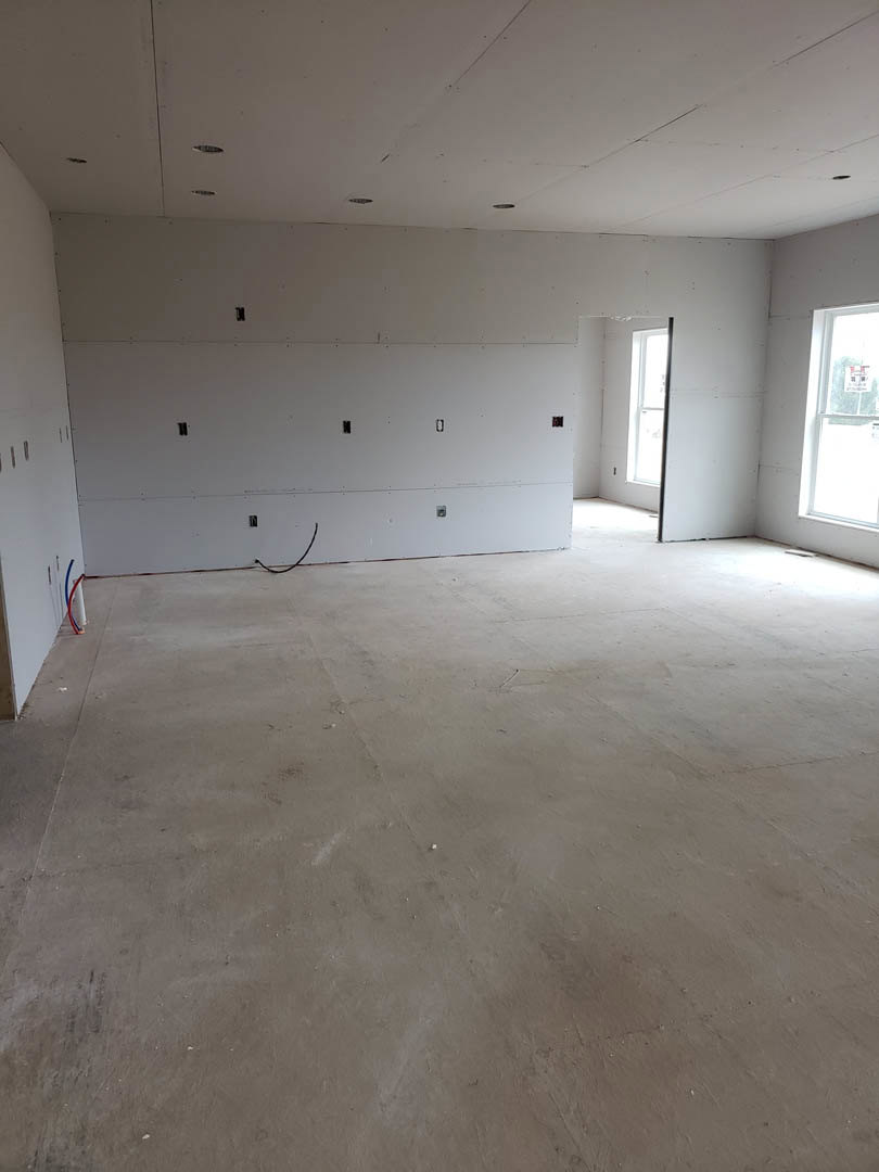 White plaster wall with drilled holes, concrete floor featuring a black wire, large windows with a sign, recessed ceiling lights, unfinished modern interior.
