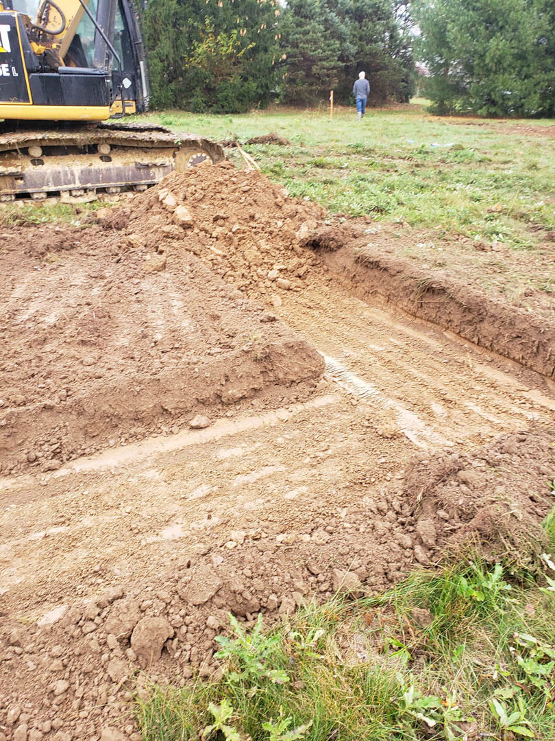 Large dirt pile on grassy field with construction equipment and tractor in the background, scattered trees and soil visible