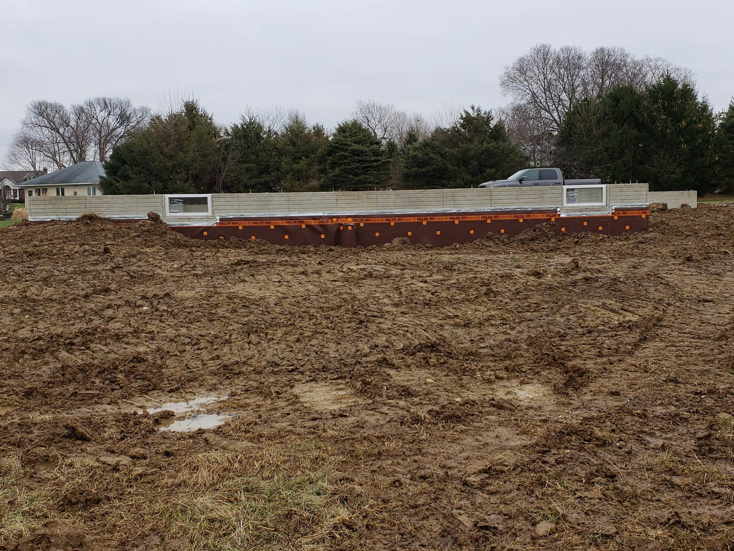 Muddy field with tire tracks, grassy patches, and a pickup truck parked near a partially constructed house; orange and black tape on a fence, trees lining the background under a