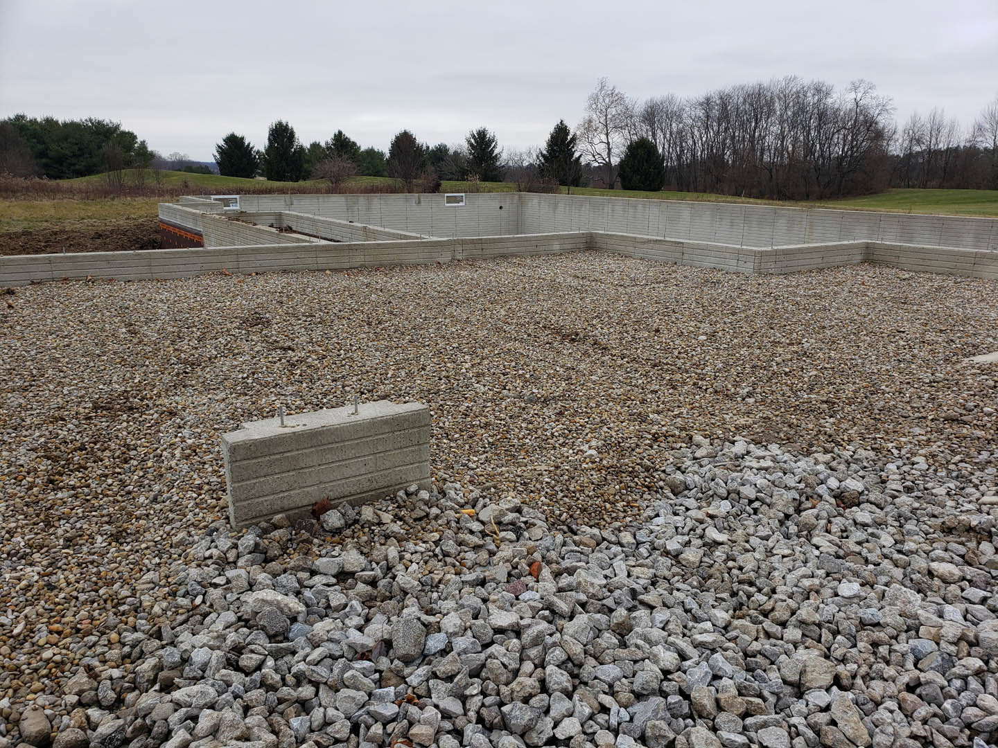 Concrete foundation surrounded by gravel and rocks, construction materials scattered on bare ground, leafy trees and blue sky in background