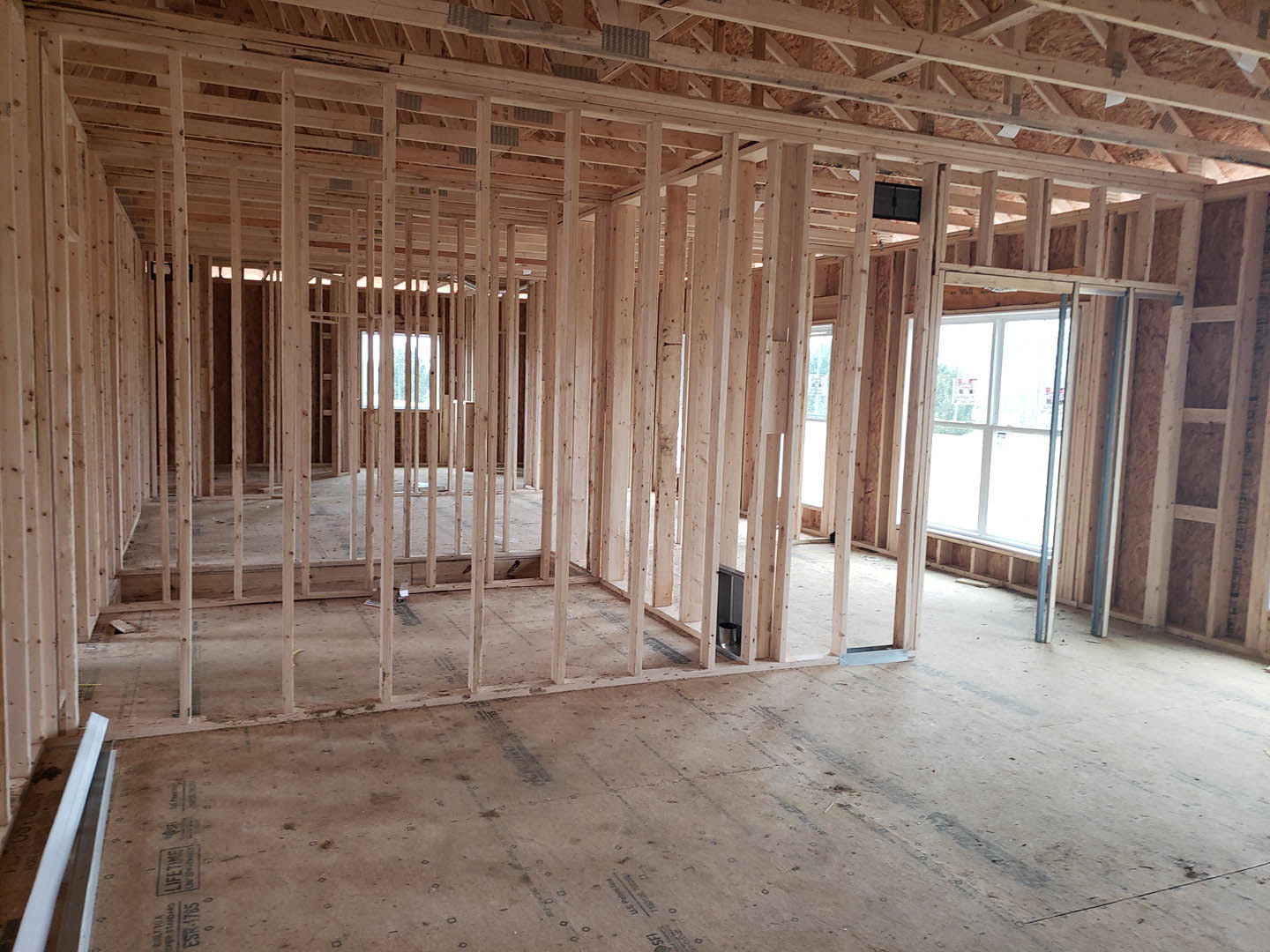 Exposed wood framing throughout unfinished room, black square object with handle near wall, wooden roof beams overhead, wood frame on floor, white door and window visible