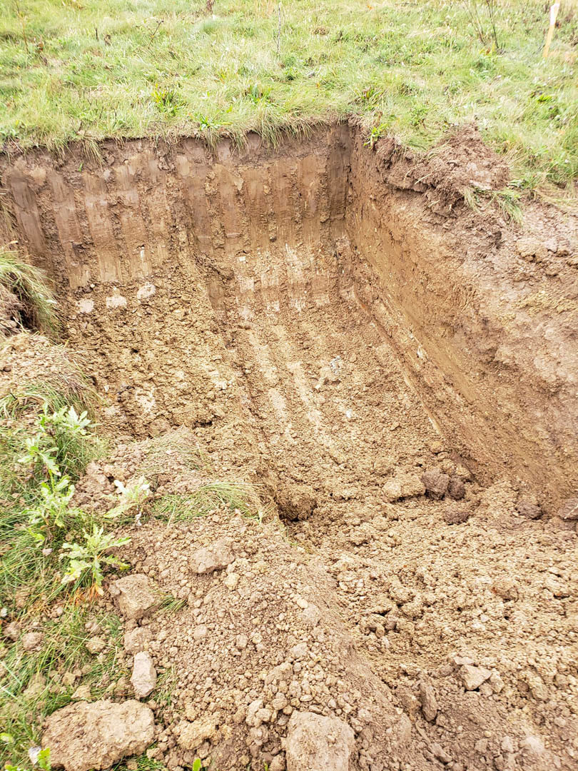 Excavated hole surrounded by loose soil and patches of grass in a residential yard