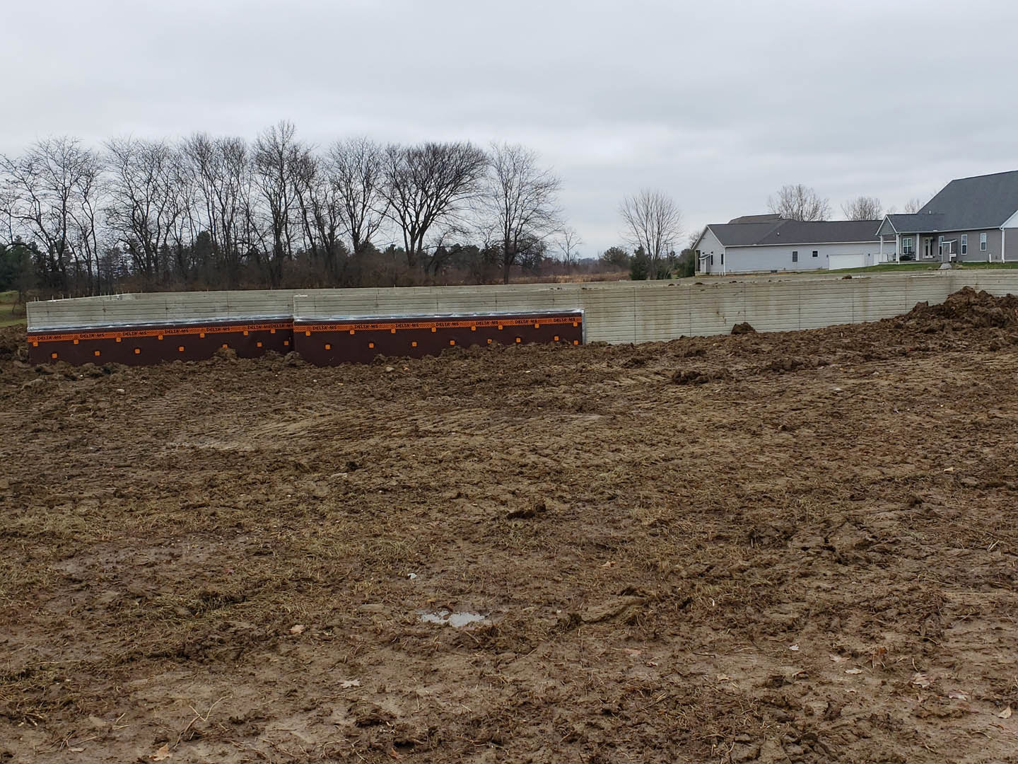 Dirt field bordered by a wooden fence, leafless trees in the background, trash cans near a house with a garage and covered porch, long wooden bench placed on bare soil