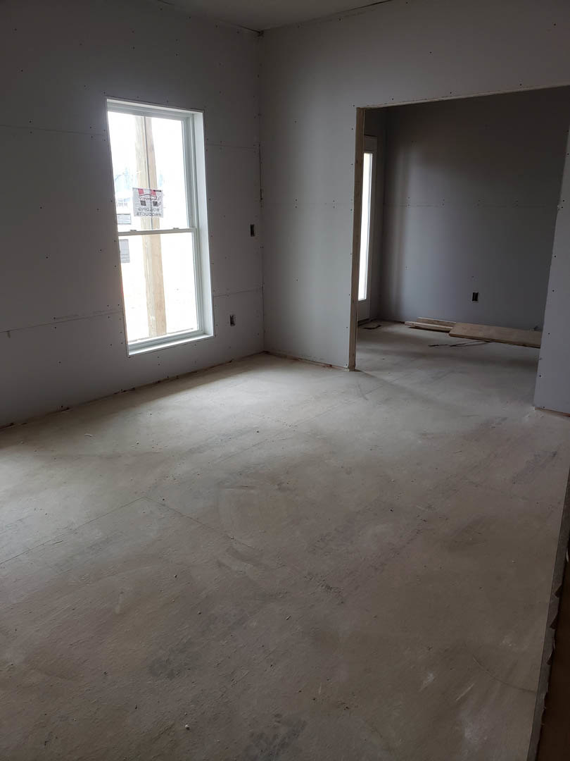 Room with light wood flooring, white plaster walls, large window with a red and white striped sign, and a wooden board resting on a table near the door.
