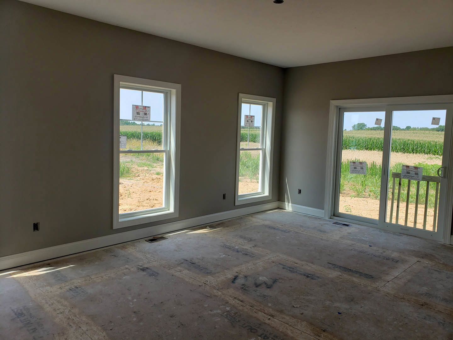 Bright room with large windows overlooking grassy field, light plaster walls, smooth flooring with black text, and signs visible on windows and lawn.