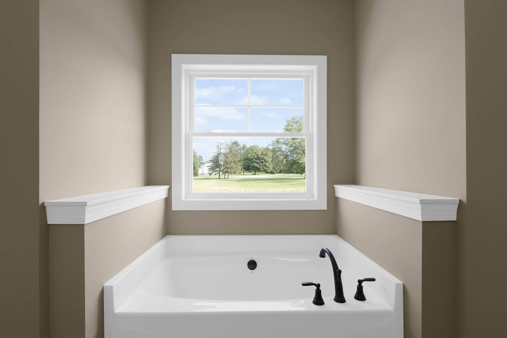 Freestanding bathtub beneath large window overlooking grassy field and trees, white wall molding, nearby sink and tap fixtures