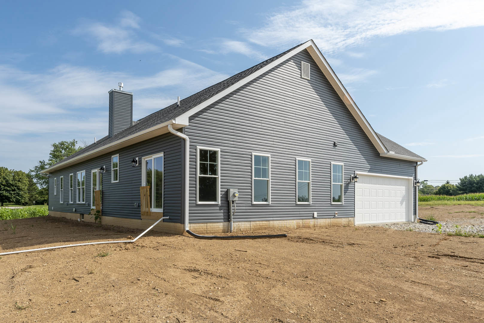 Two-story house with white garage door, brick chimney, white-framed windows, wooden porch chair, and black pipe on dirt beside the exterior.