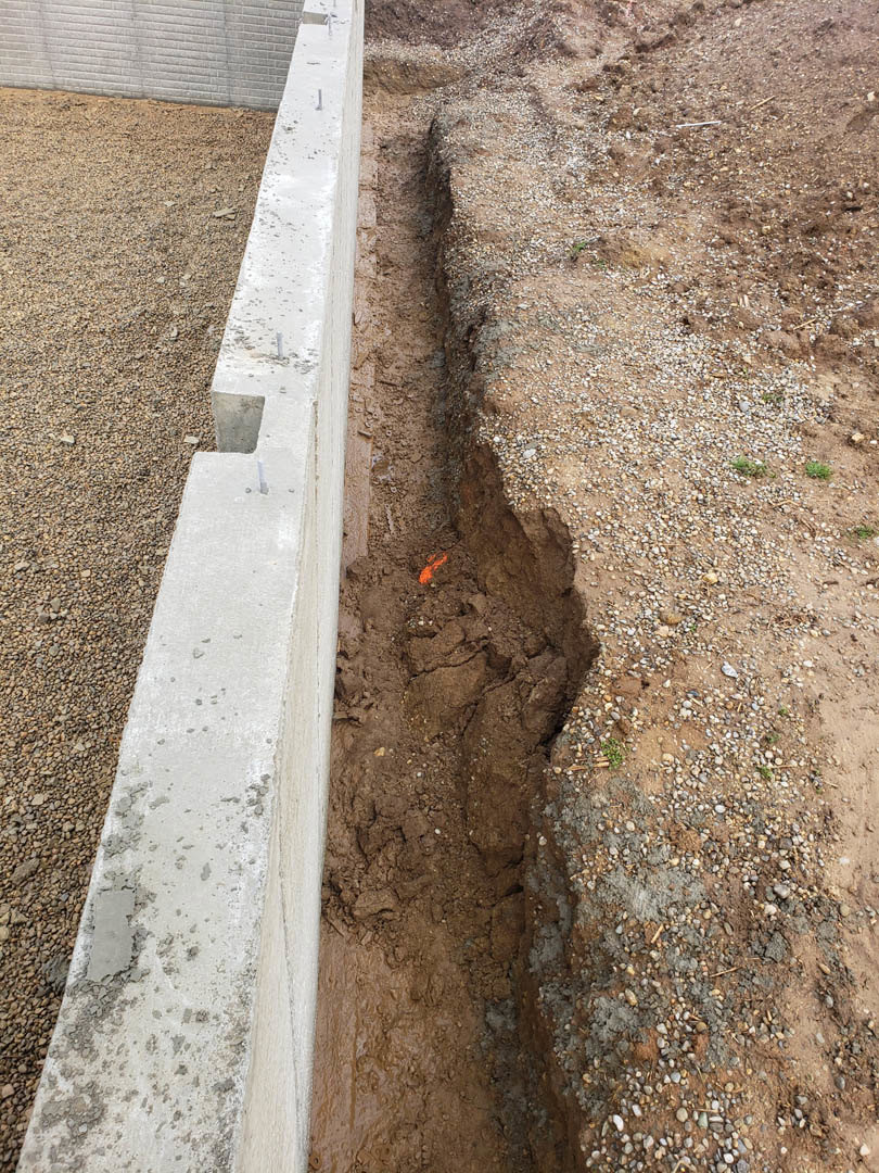 Wide crack running through rocky dirt ground beside concrete and white brick walls with scattered gravel