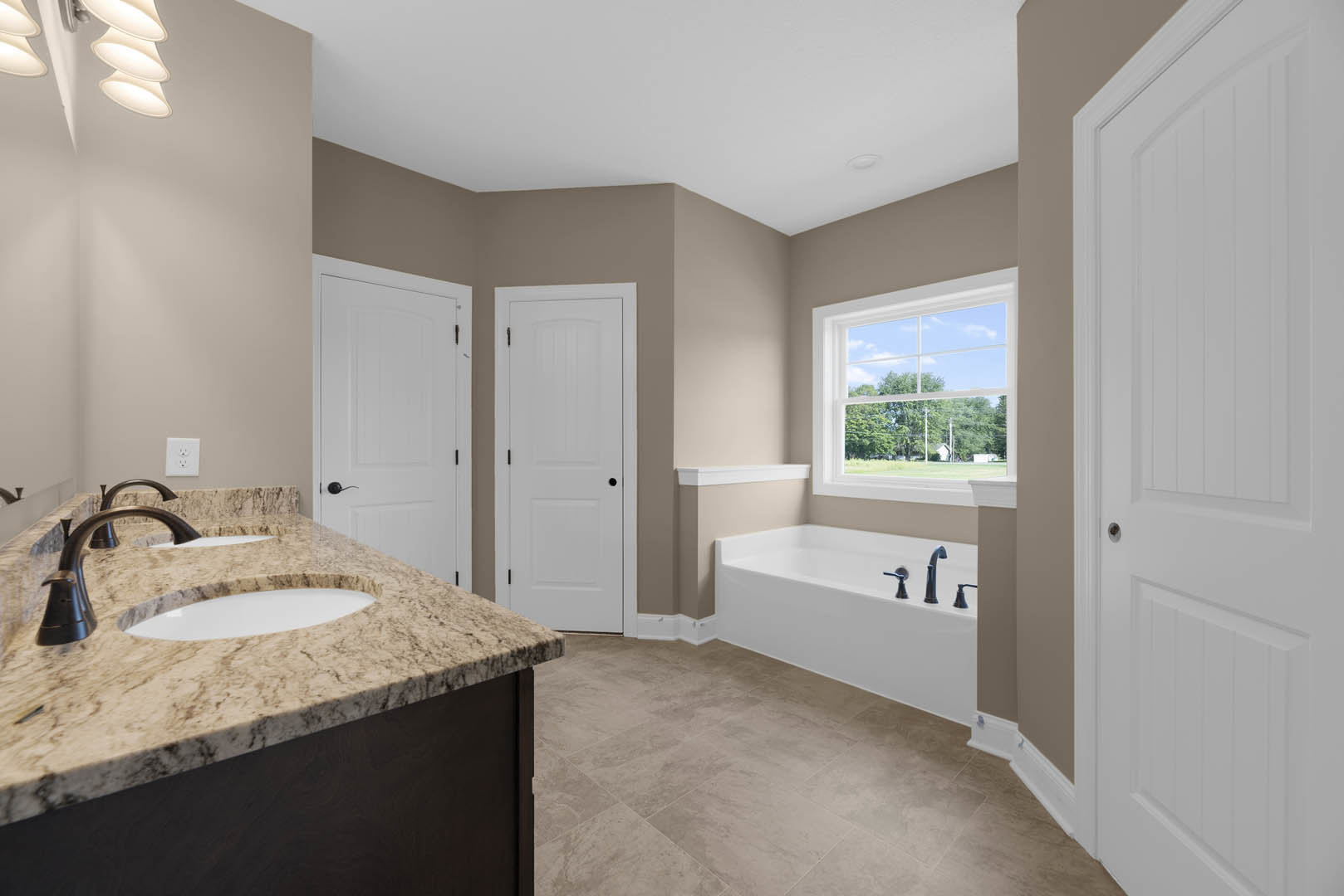 Bathroom with marble countertop and tub, window overlooking field and trees, white door with black knobs, double outlet, sink with chrome faucets, white cabinetry
