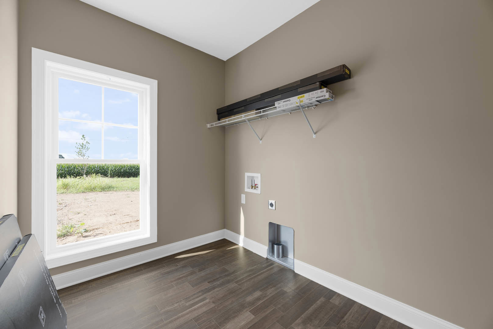 Bright room with laminate wood flooring, white plaster walls, large window overlooking a grassy field, built-in shelf holding storage boxes and a silver can, white ceiling with a