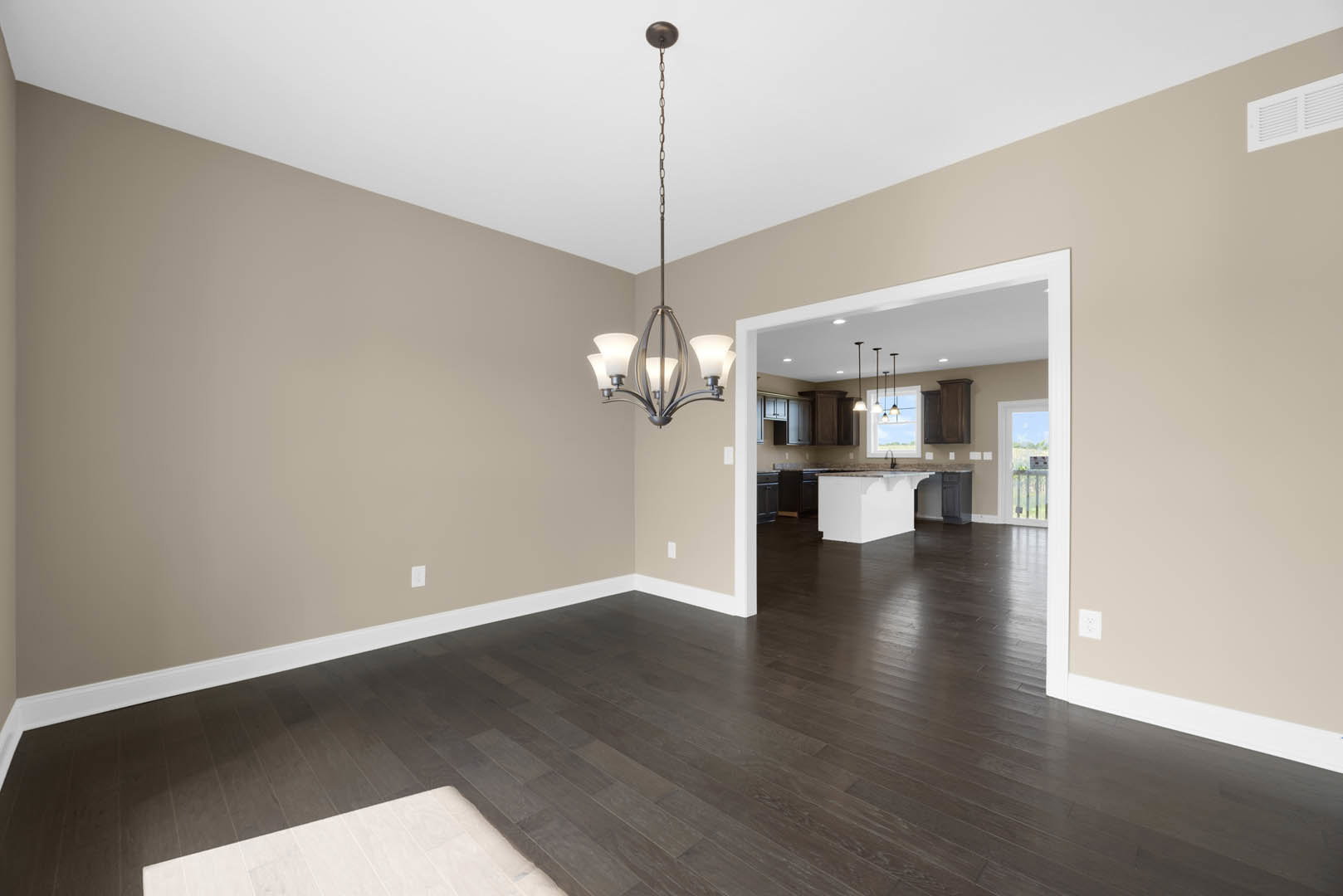 Open kitchen and dining area with dark wood flooring, white cabinetry, marble countertops, and a modern chandelier hanging above a dining table; brown wooden door and white walls