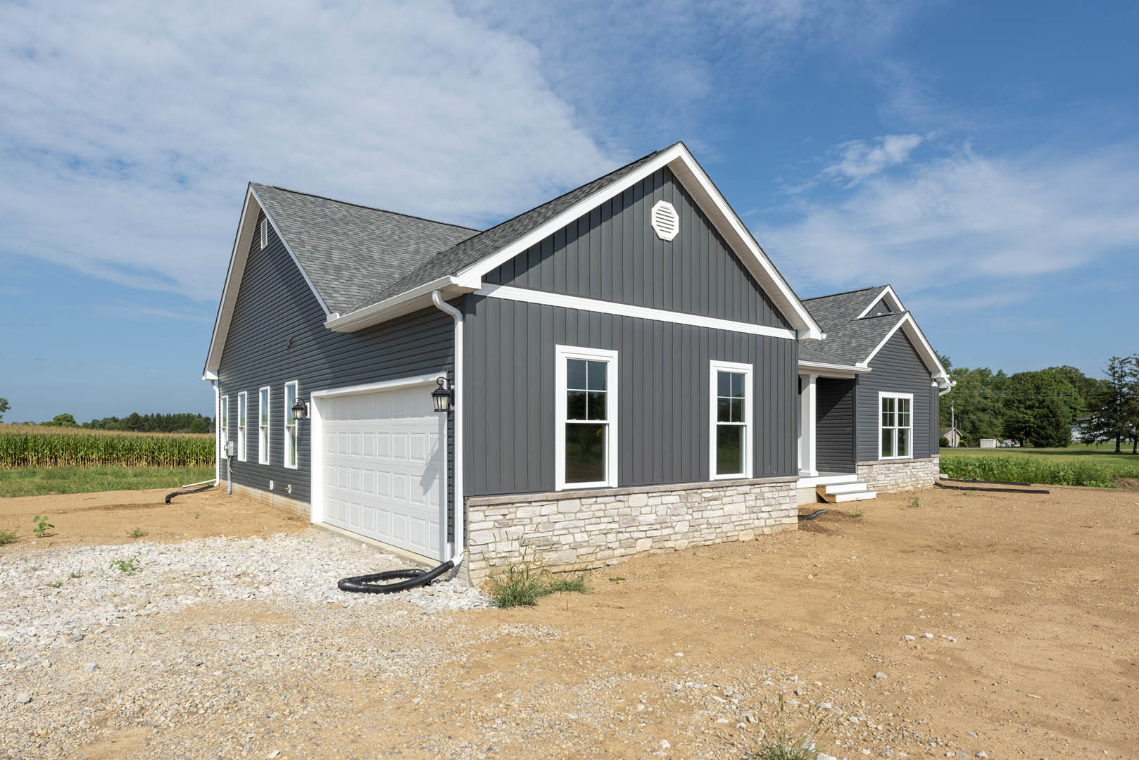 Grey siding house with white garage door, exterior light above garage, drainage pipe along wall, close-up window, gabled roof under partly cloudy sky.