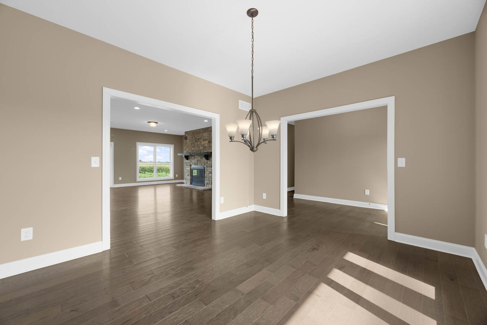 Hardwood floored living room with brick accent wall, fireplace, large window overlooking greenery, chandelier suspended from ceiling, and white electrical outlet on plaster wall
