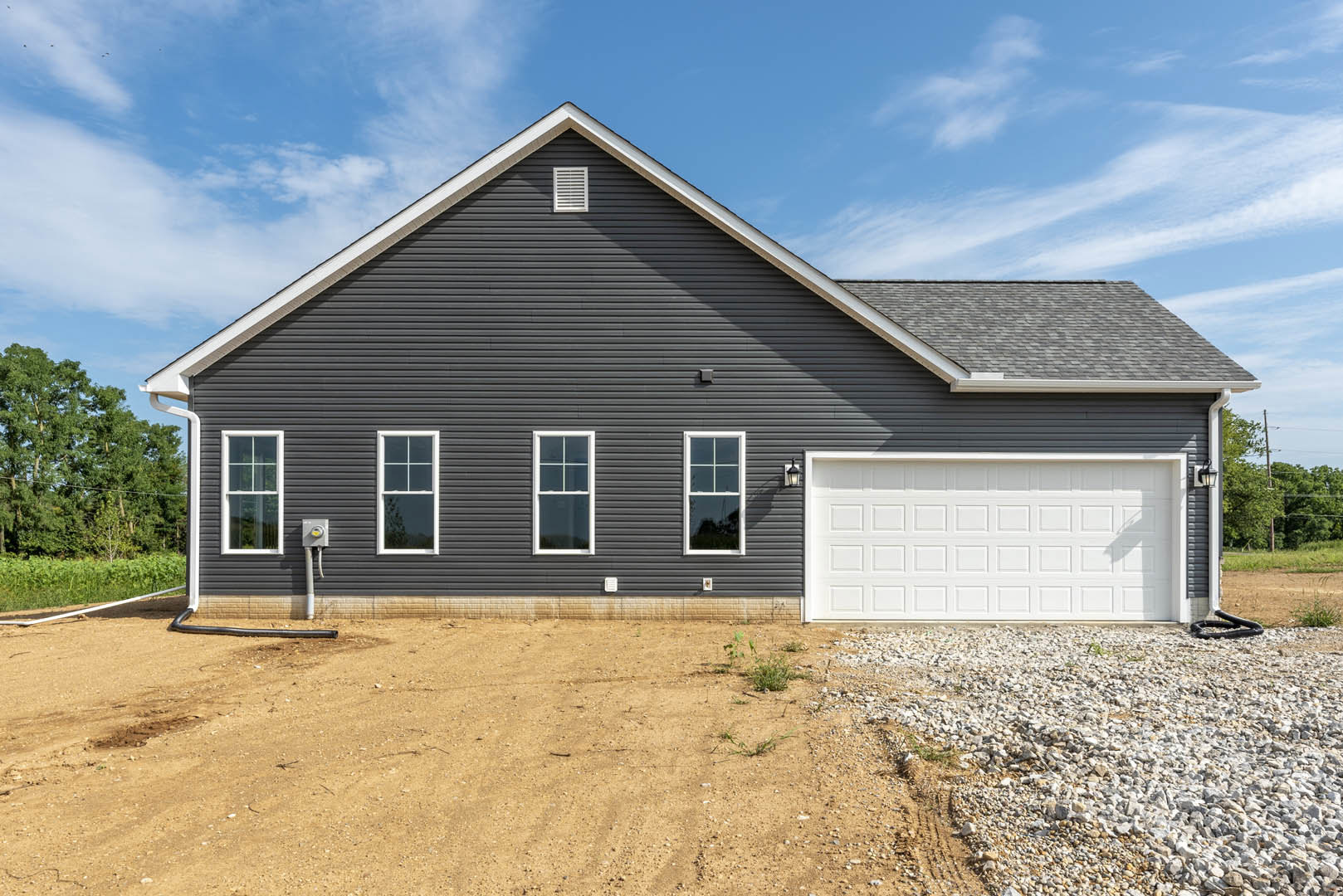 White house with attached garage, white-framed windows, light siding, and green trees in background; garage door closed, pile of rocks and dirt in foreground.